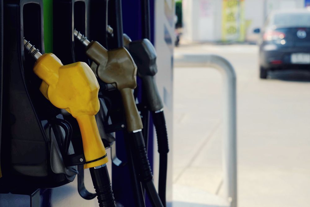 Gas pump nozzles, yellow, green and black, at a gas station with a car in the background.