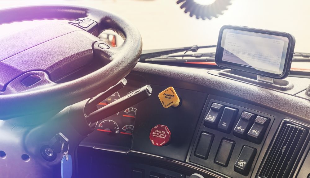 Interior of a semi-truck cab: steering wheel, dashboard with buttons, cell phone, and two red and yellow warning stickers.
