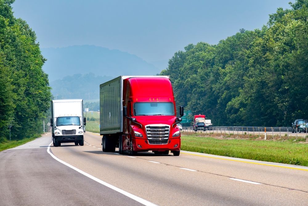 Red semi-truck and white truck on a highway flanked by trees, sunny day.