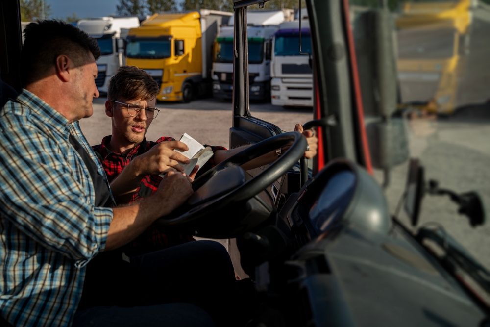 Two people in a truck cab, one teaching the other. Trucks in background.
