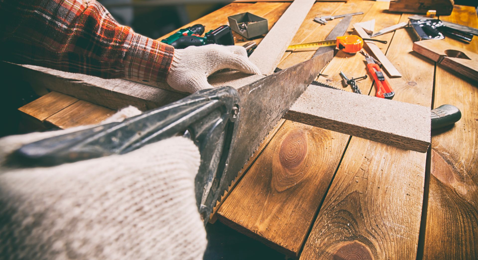 A man wearing work gloves is using a saw to cut through a wooden plank placed on a workbench.