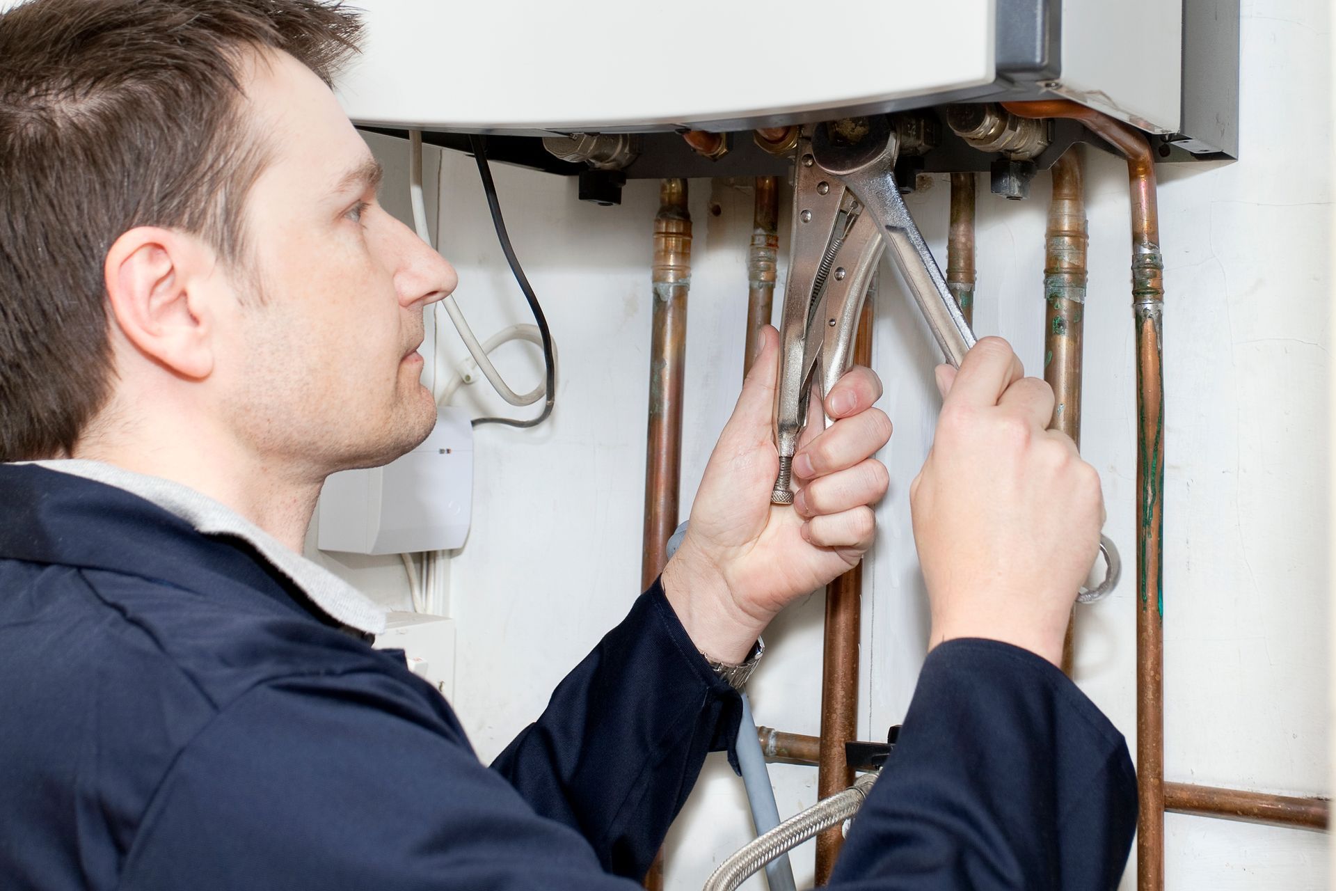 Man in a dark blue work suit, repairing copper pipes with pliers near a white appliance.