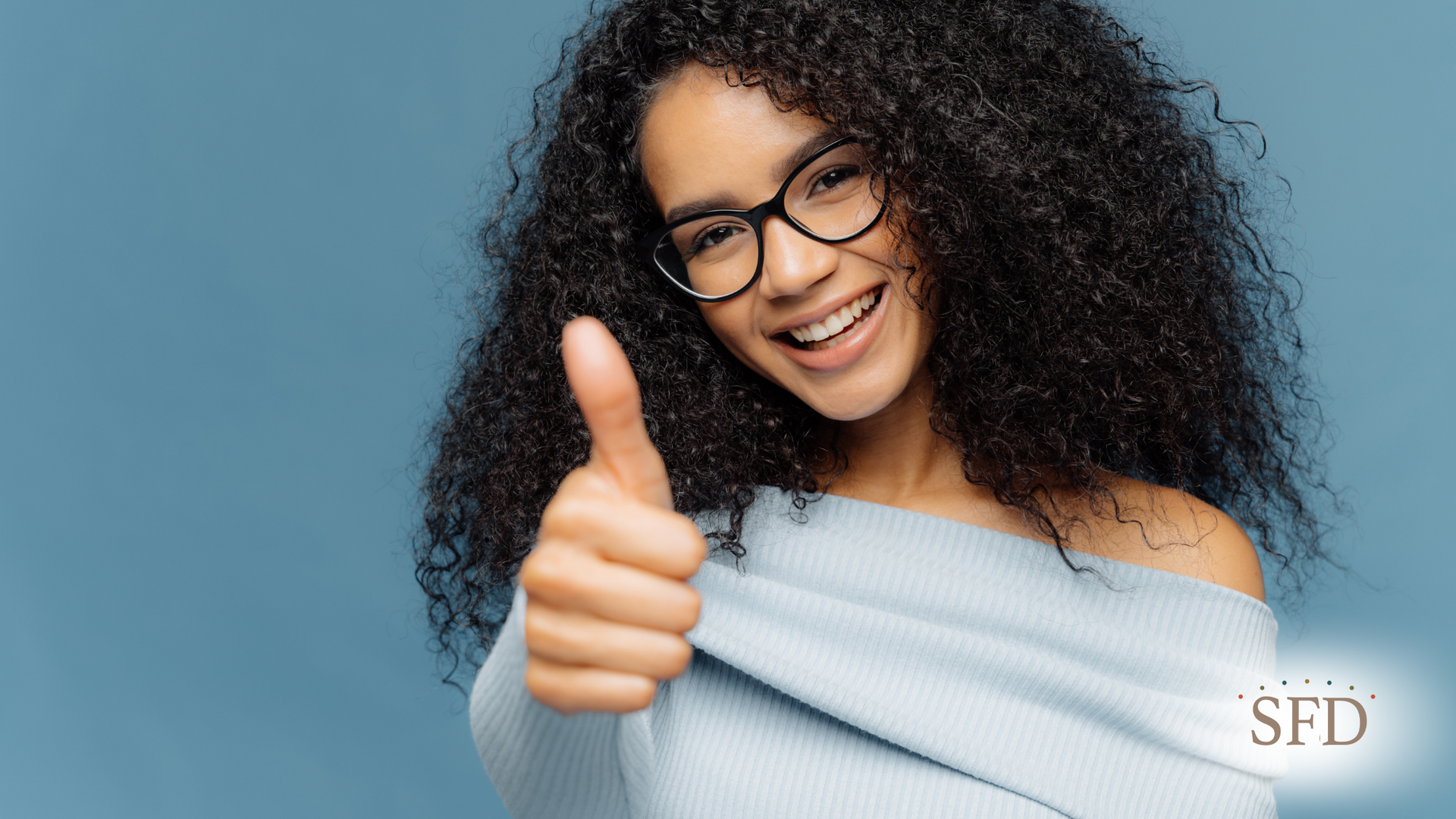 A woman with curly hair and glasses is smiling and giving a thumbs up.