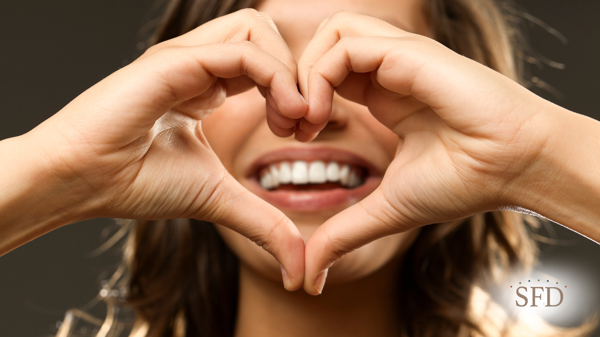 Woman smiles, forming a heart shape with hands, revealing white teeth.