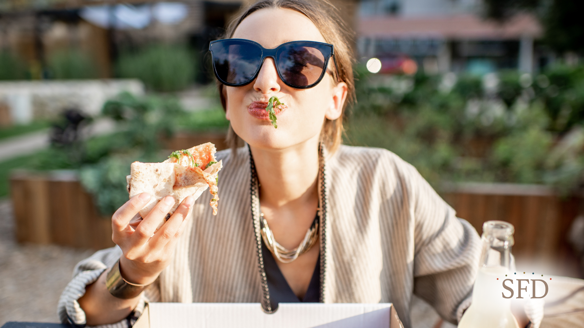 Woman in sunglasses eating pizza outdoors, smiling.