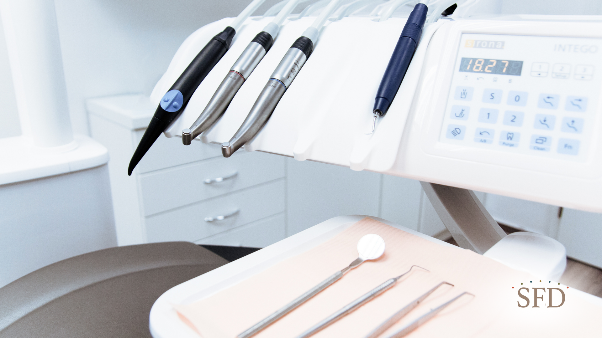Dental tools on a tray, with dental chair, and equipment in the background. White and silver tones.