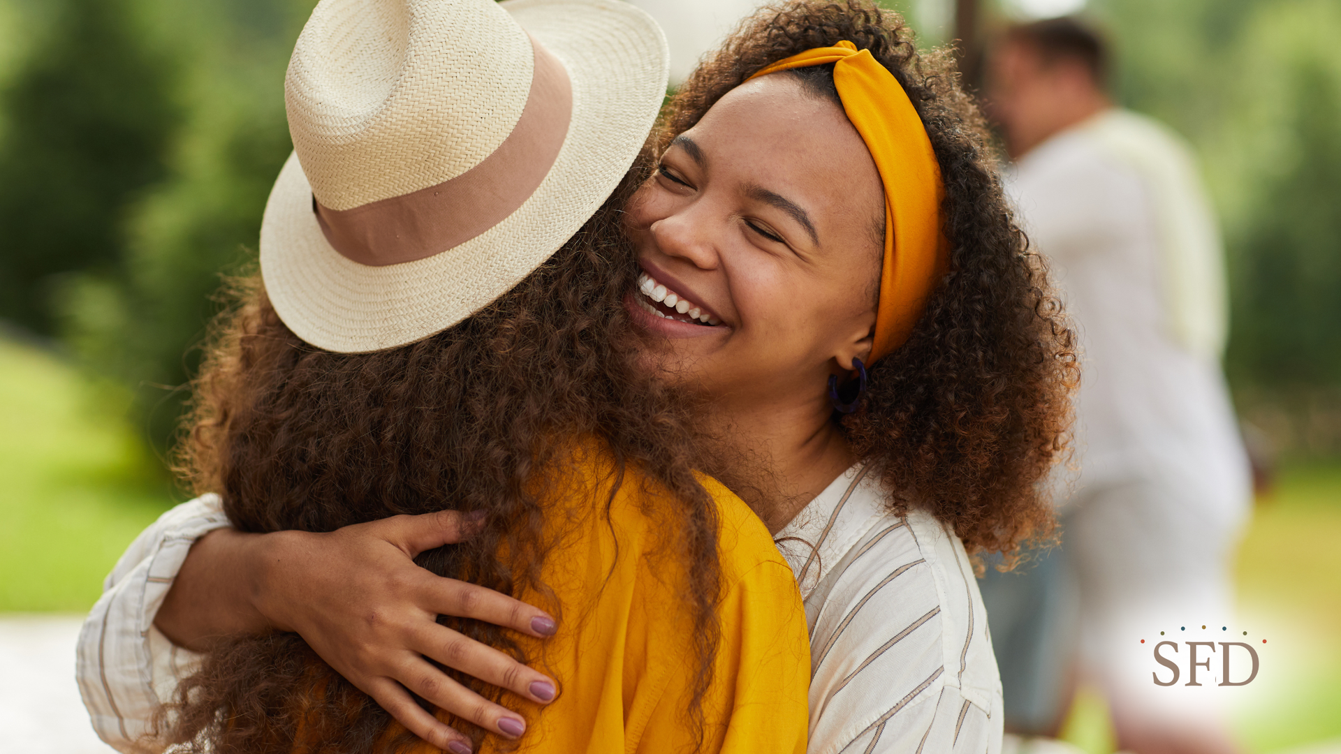 Two women embracing outdoors, smiling. One wearing a hat, the other a yellow headband.