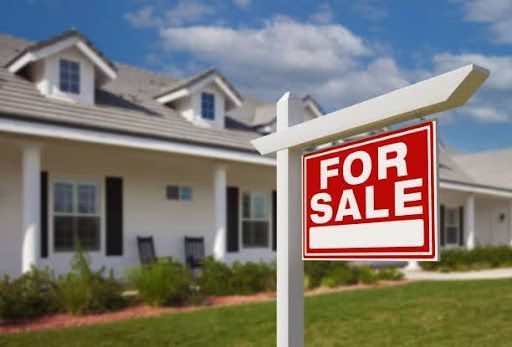 A red For Sale sign in front of a white home with a lawn and blue sky.