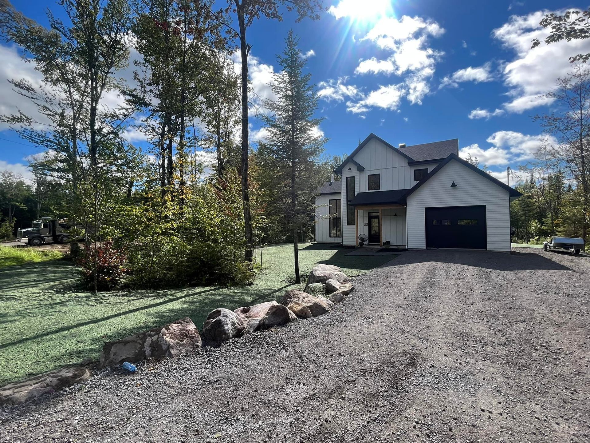 Une maison blanche avec une porte de garage noire est entourée d'arbres et de gravier.