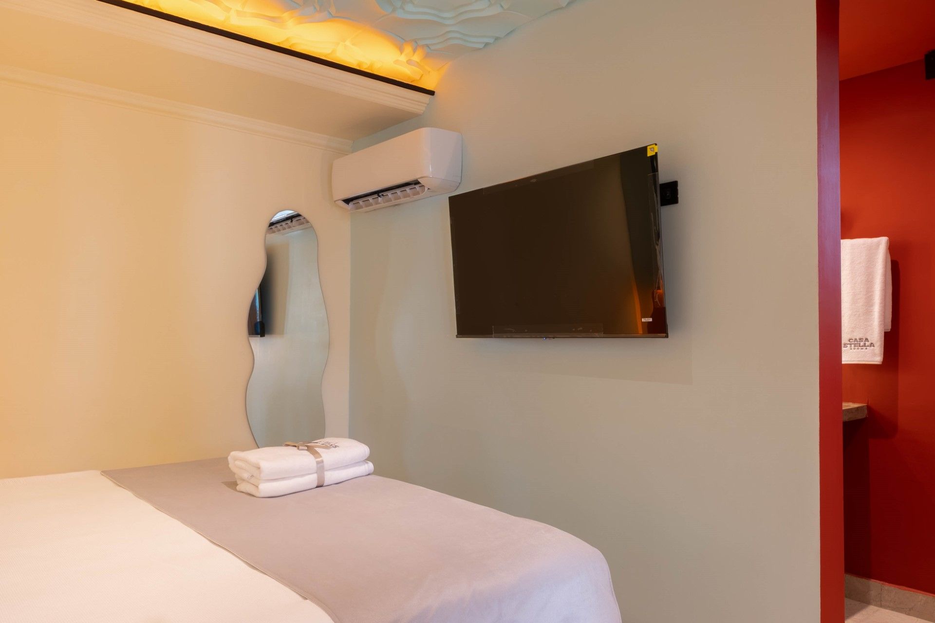 Dormitorio con cama, televisor de pared, aire acondicionado y toallas. Puerta roja y espejo en la pared.