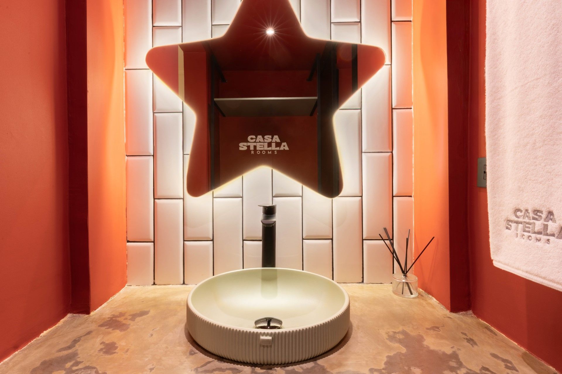 Baño con espejo en forma de estrella, lavabo redondo y toalla con el logo de CASA STELLA. Paredes rojas y azulejos blancos.