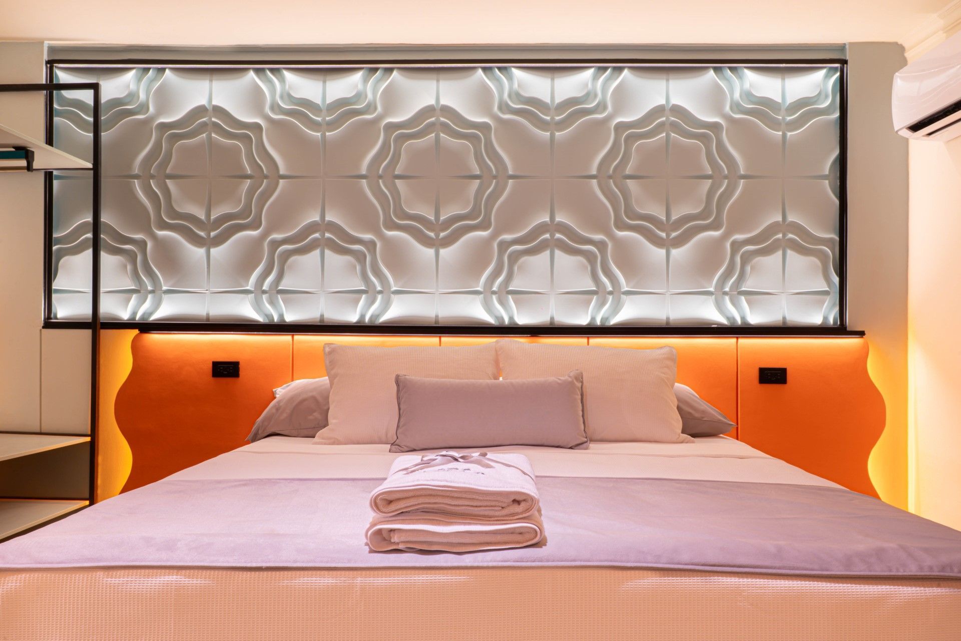 Dormitorio con cabecero estampado iluminado por luces naranjas, encima de una cama con toallas dobladas.