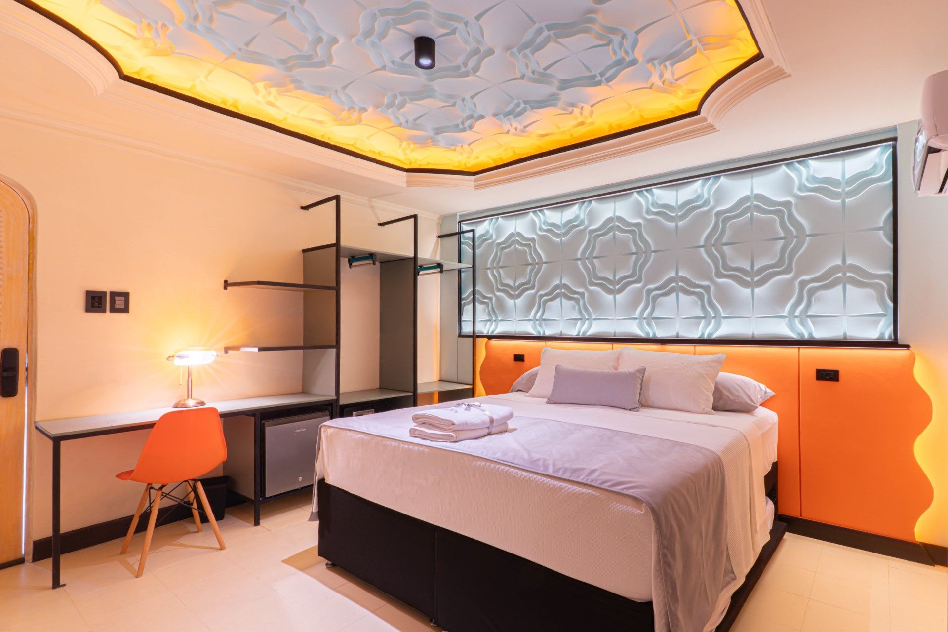 Habitación de hotel con cama, escritorio, armario abierto, techo estampado y panel de pared decorativo.