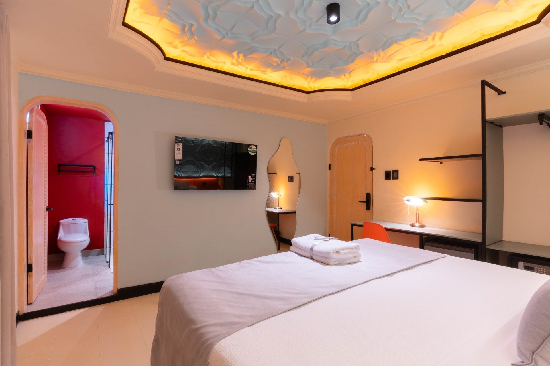 Dormitorio con cama, televisor, escritorio y puerta de baño. Techo con iluminación decorativa y diseño.