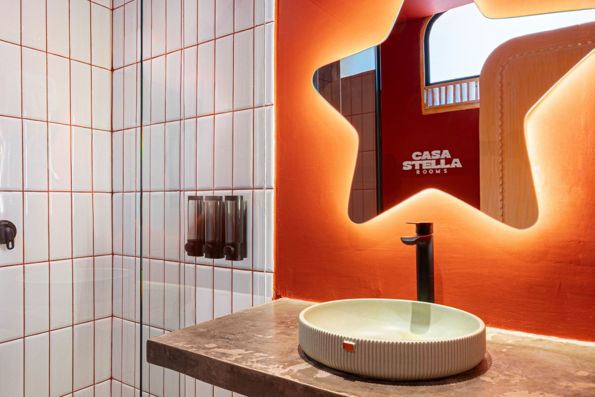 Baño con ducha de azulejos blancos, pared naranja con espejo en forma de estrella y lavabo sobre encimera.