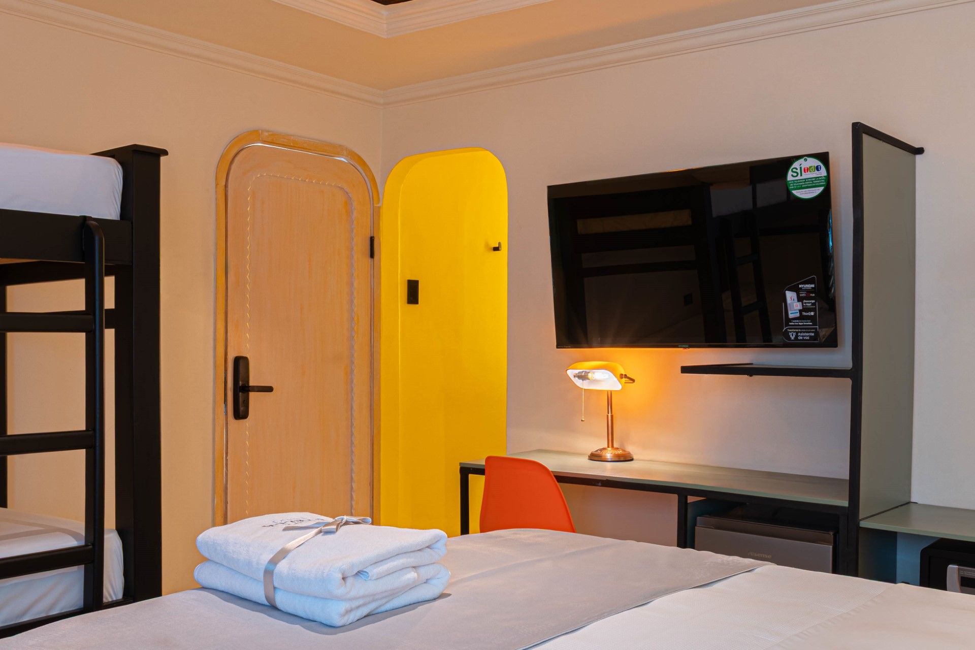 Literas, toallas dobladas y una cama en una habitación con TV y una puerta de color amarillo brillante.