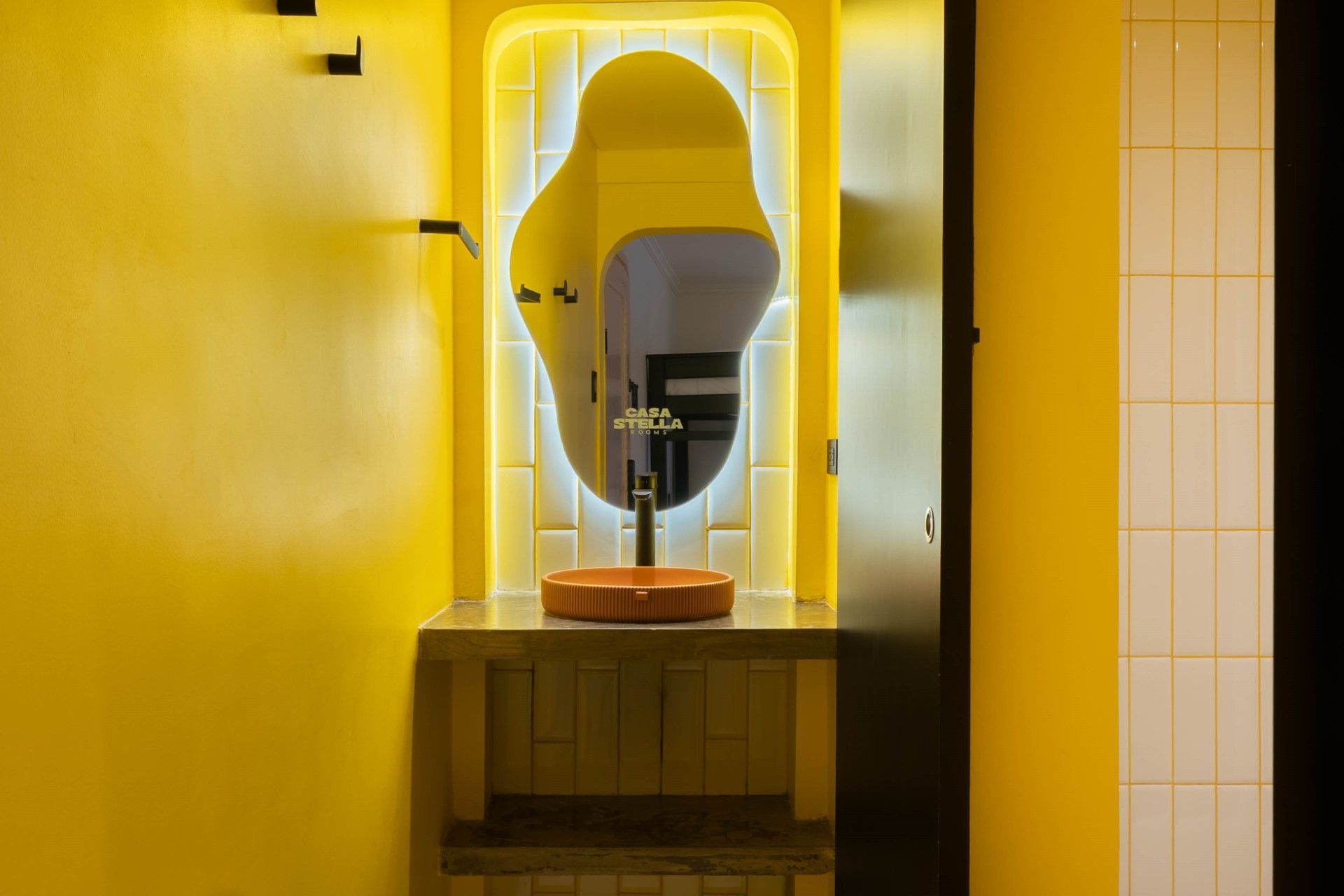 Baño amarillo con espejo de forma orgánica, lavabo de madera y nicho retroiluminado.