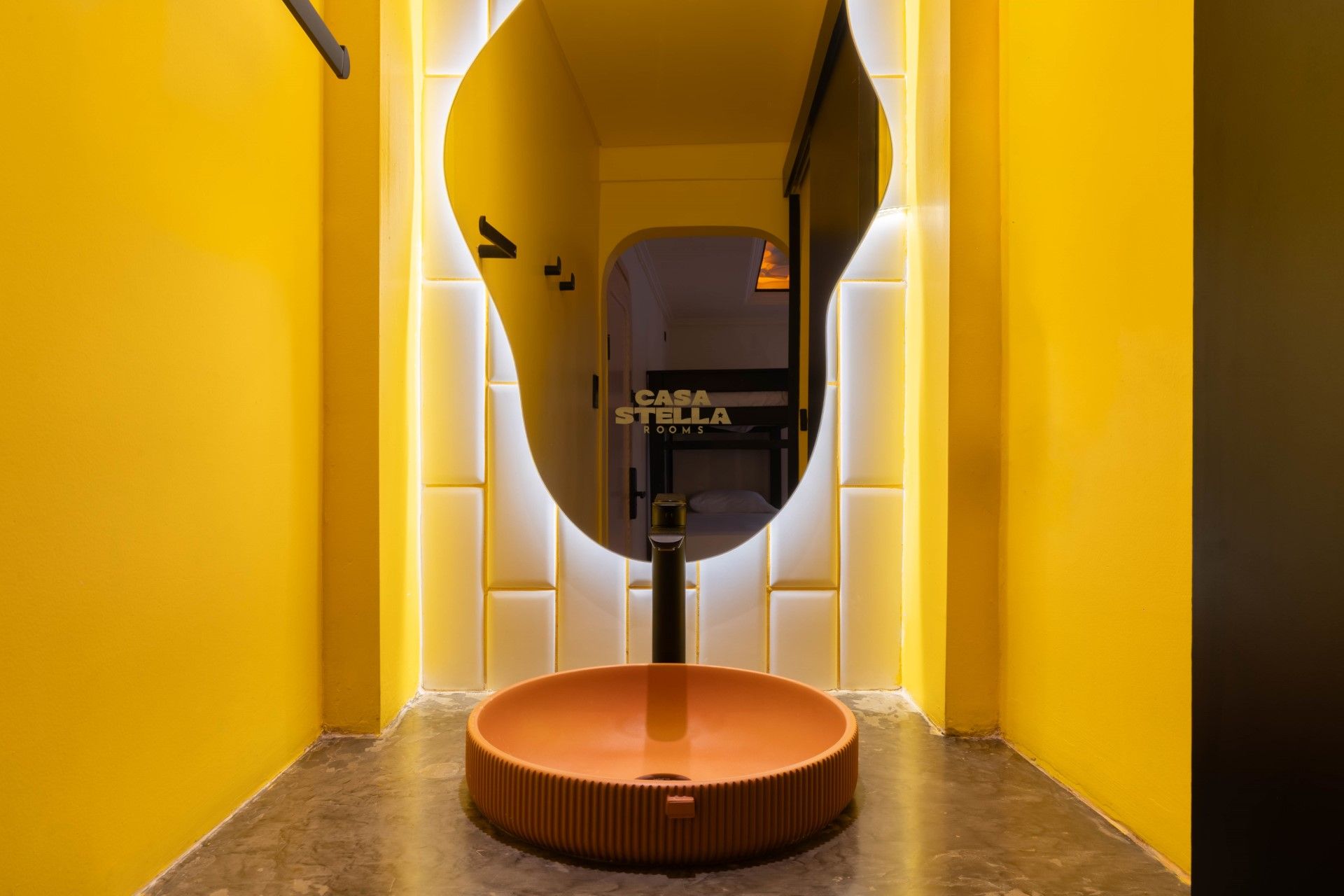 Baño amarillo con espejo de forma orgánica, lavabo de terracota y pared de azulejos.