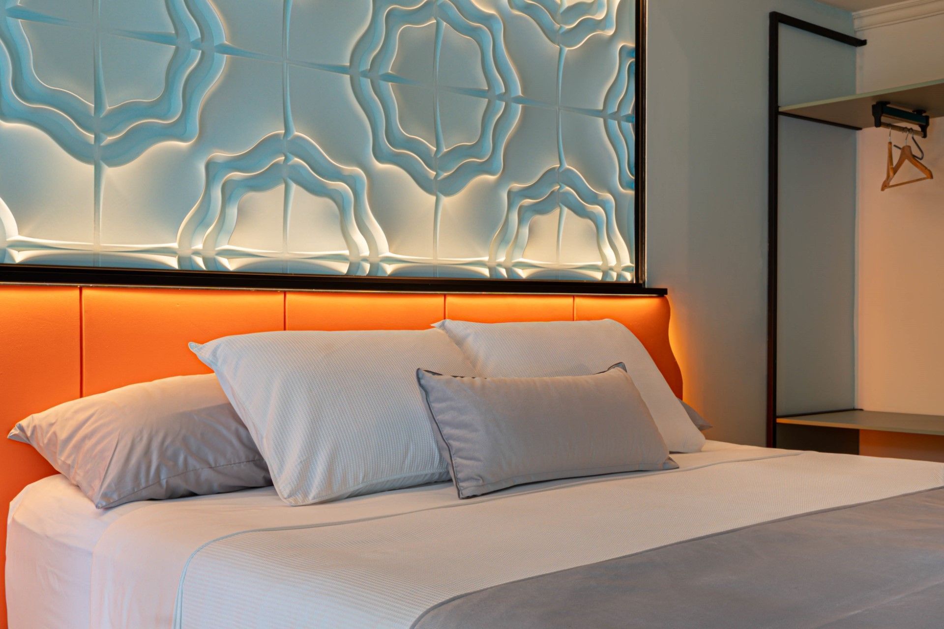 Dormitorio con cabecera naranja, ropa de cama blanca, almohadas grises y un panel de pared decorativo iluminado en azul.
