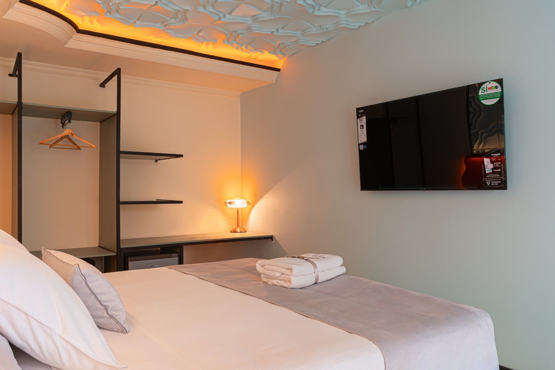 Habitación de hotel: cama, televisor, armario, lámpara y techo texturizado. Colores neutros, iluminación cálida.