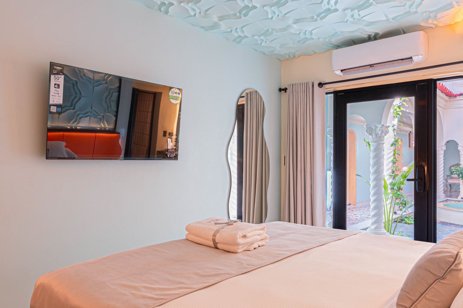 Dormitorio con cama, TV de pared, espejo de cuerpo entero y puertas que dan a un patio.