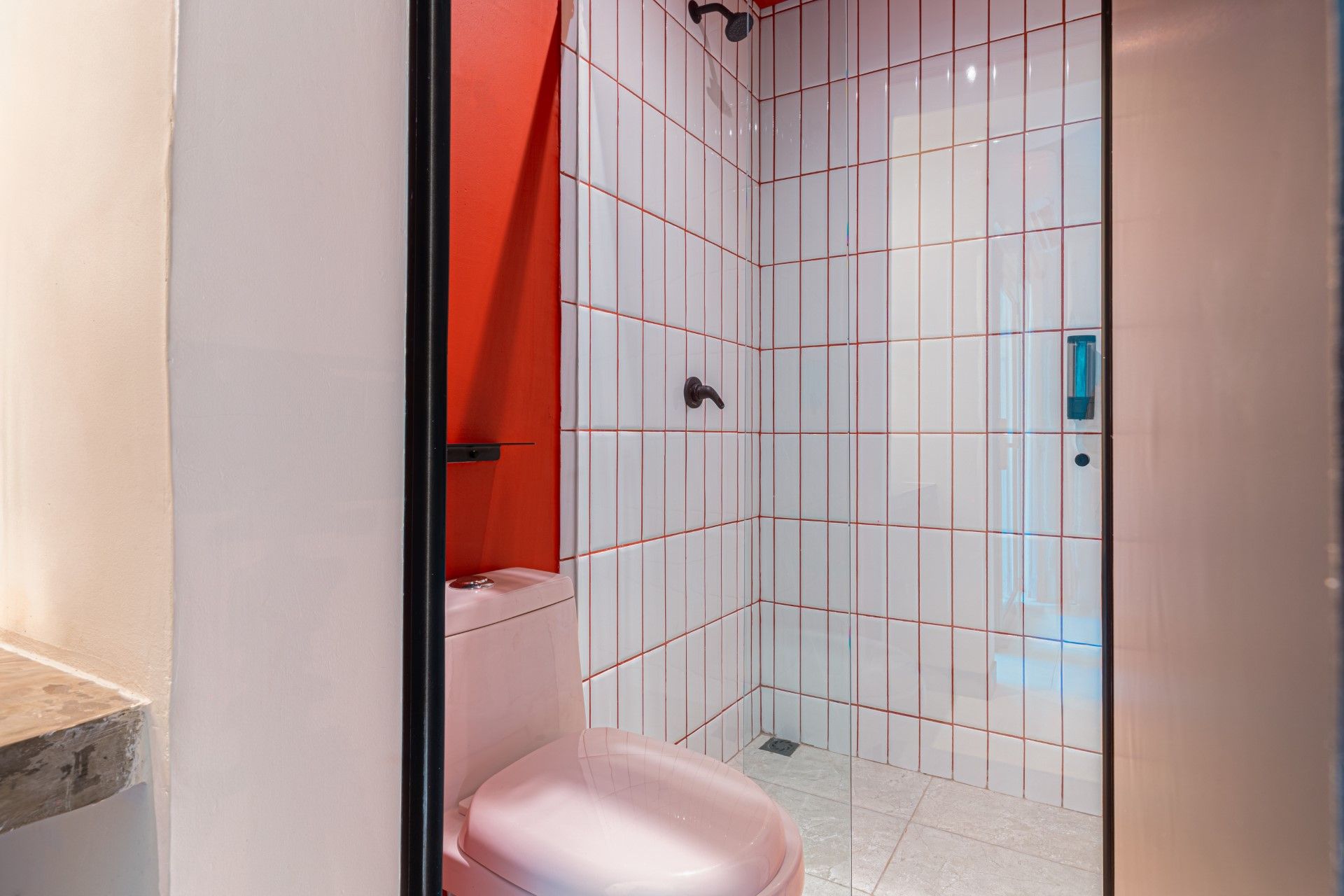 Pequeño baño con ducha de azulejos blancos, inodoro rosa y pared con acento rojo.