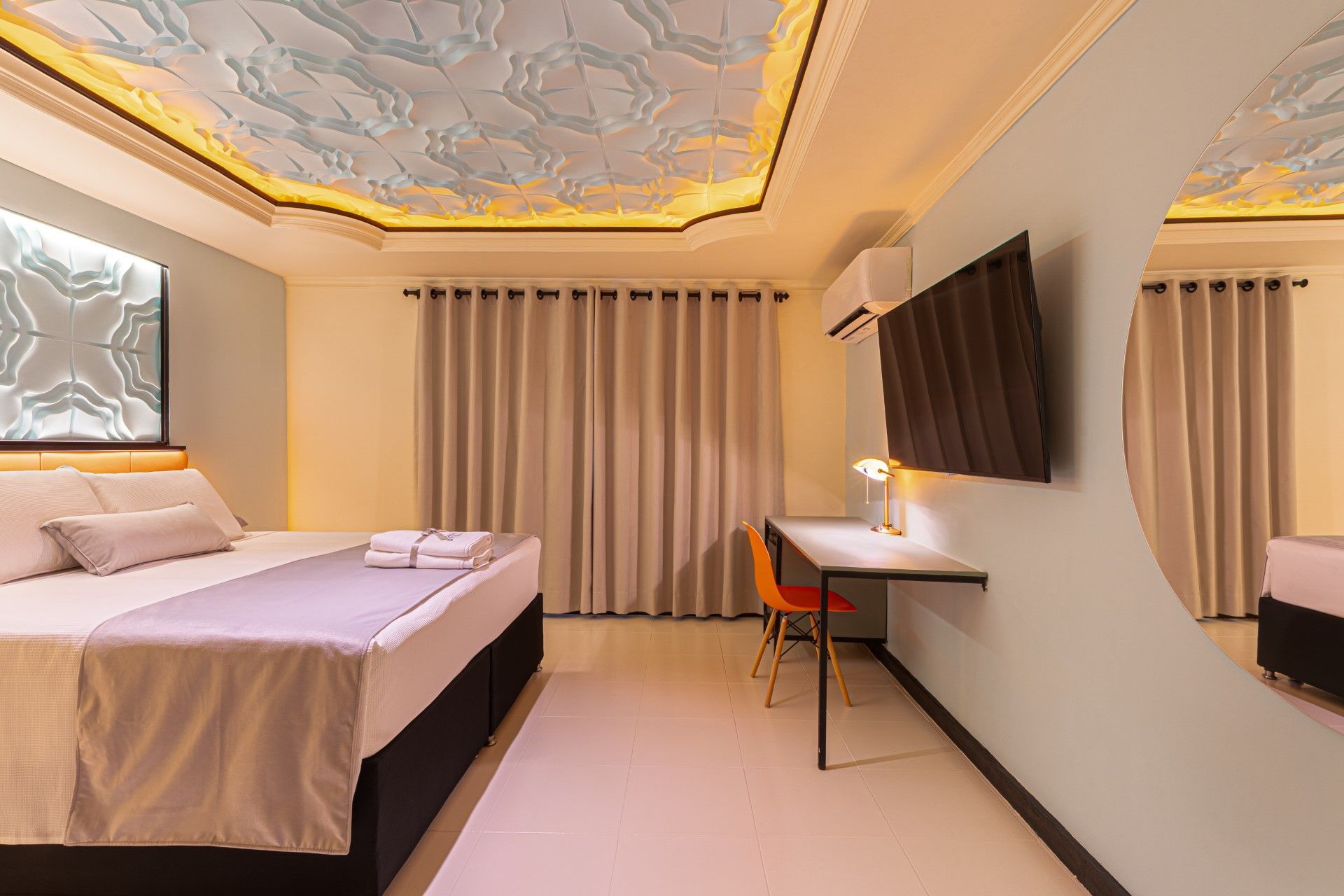 Habitación de hotel con cama, escritorio, televisor y techo decorativo. Tonos beige, azul claro y blanco.