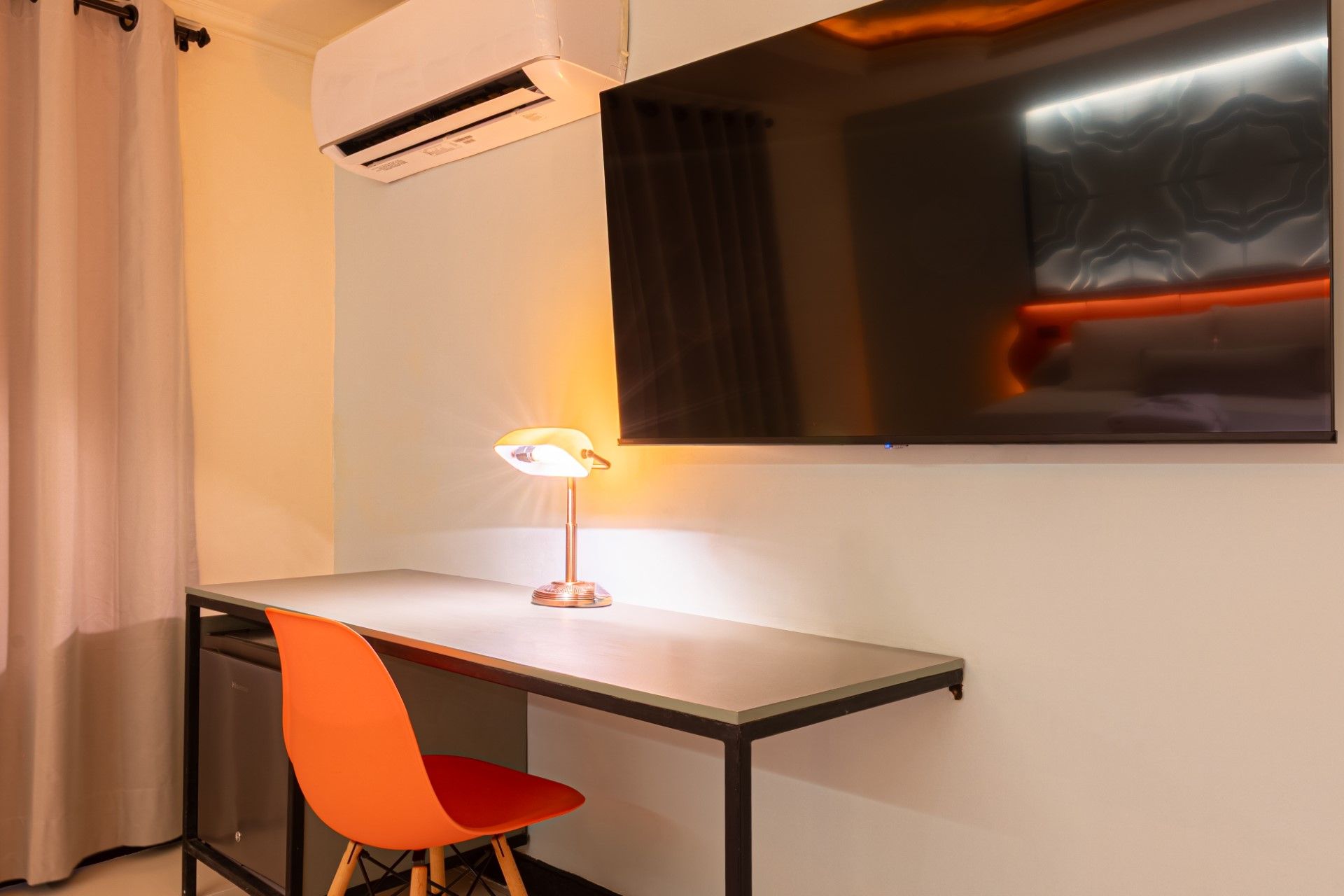 Escritorio con silla naranja, lámpara y televisor en la pared. Aire acondicionado y cortina visibles.