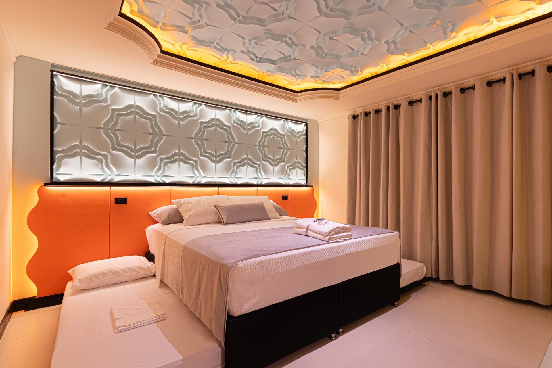 Dormitorio con cama, cabecero, techo decorativo y cortinas; iluminado con cálida luz naranja y blanca.