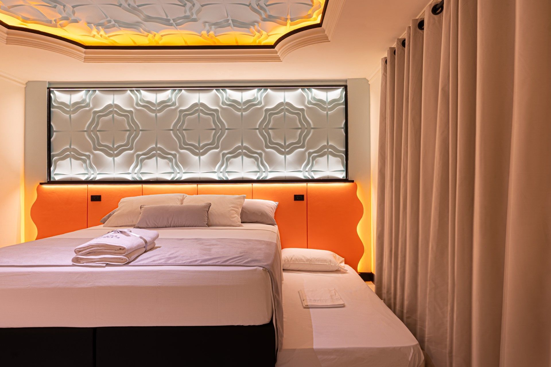 Dormitorio con cabecero naranja, panel decorativo en la pared, cama con sábanas y sofá cama. Cortinas beige.