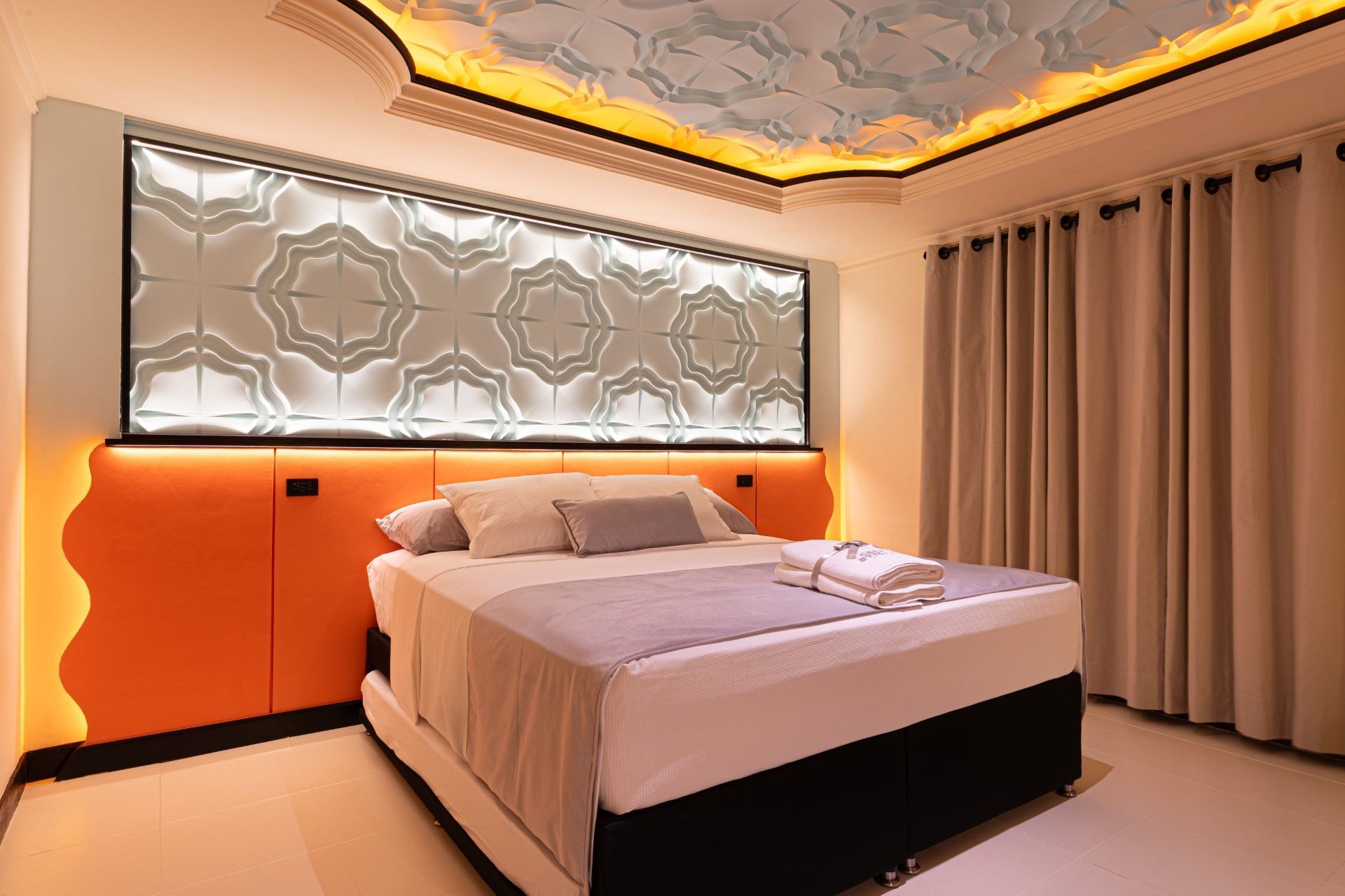 Dormitorio con cabecero naranja, panel decorativo en la pared y techo ornamentado. La cama está hecha con toallas.