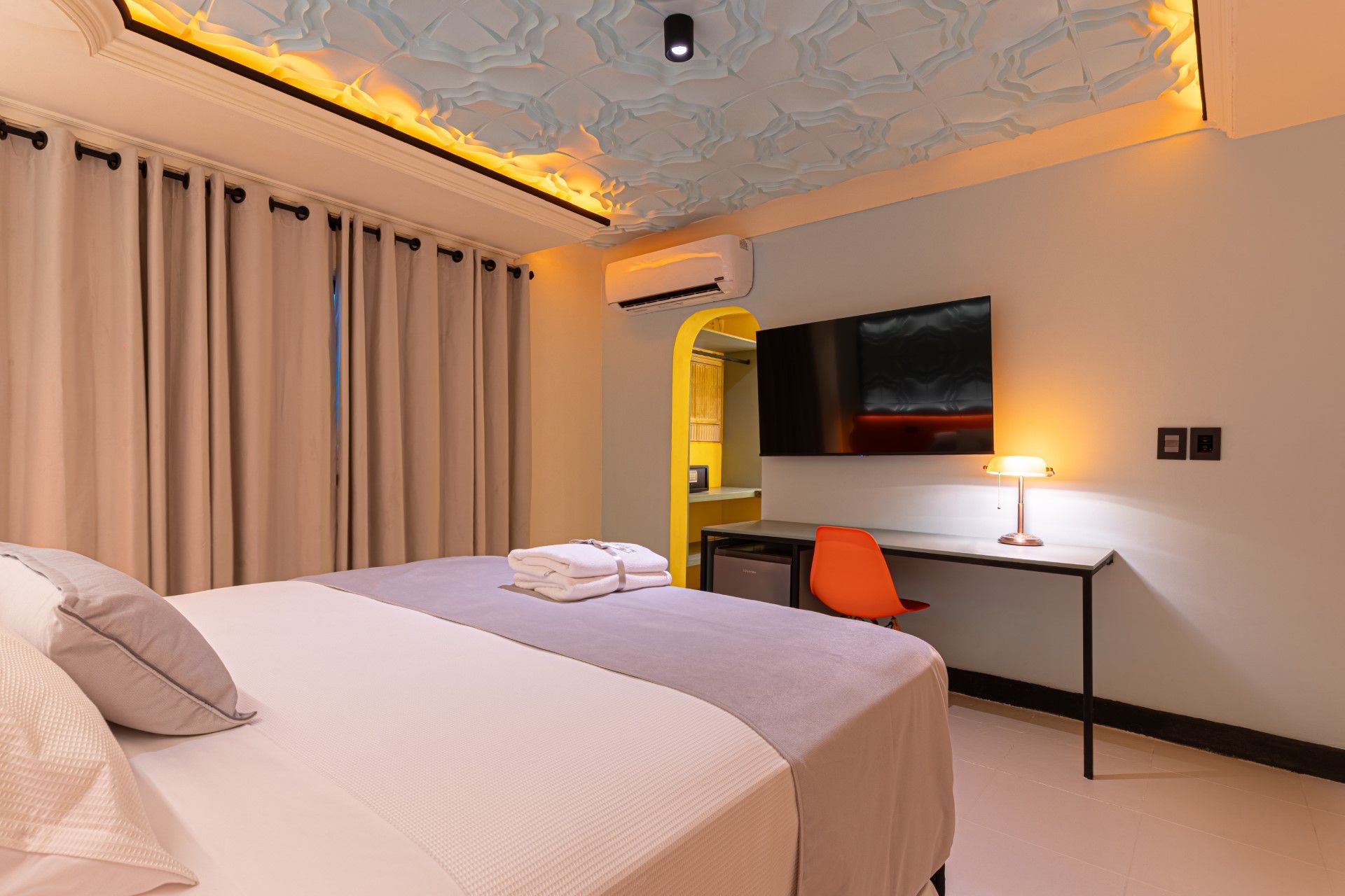 Dormitorio con cama grande, televisor, escritorio y puerta arqueada. El techo presenta iluminación cálida.