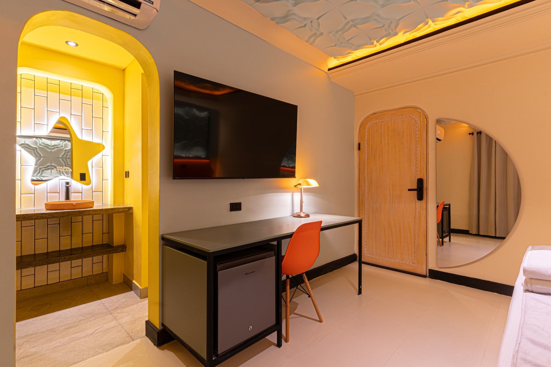 Habitación de hotel con escritorio, TV, nevera y puerta que da a un baño con espejo en forma de estrella.