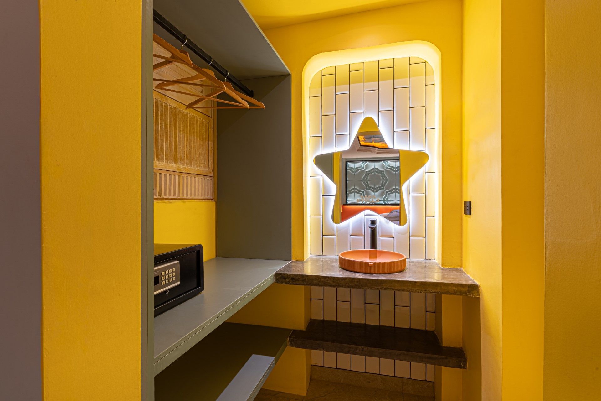 Rincón de habitación amarillo con armario, caja fuerte y espejo iluminado en forma de estrella sobre un mostrador.