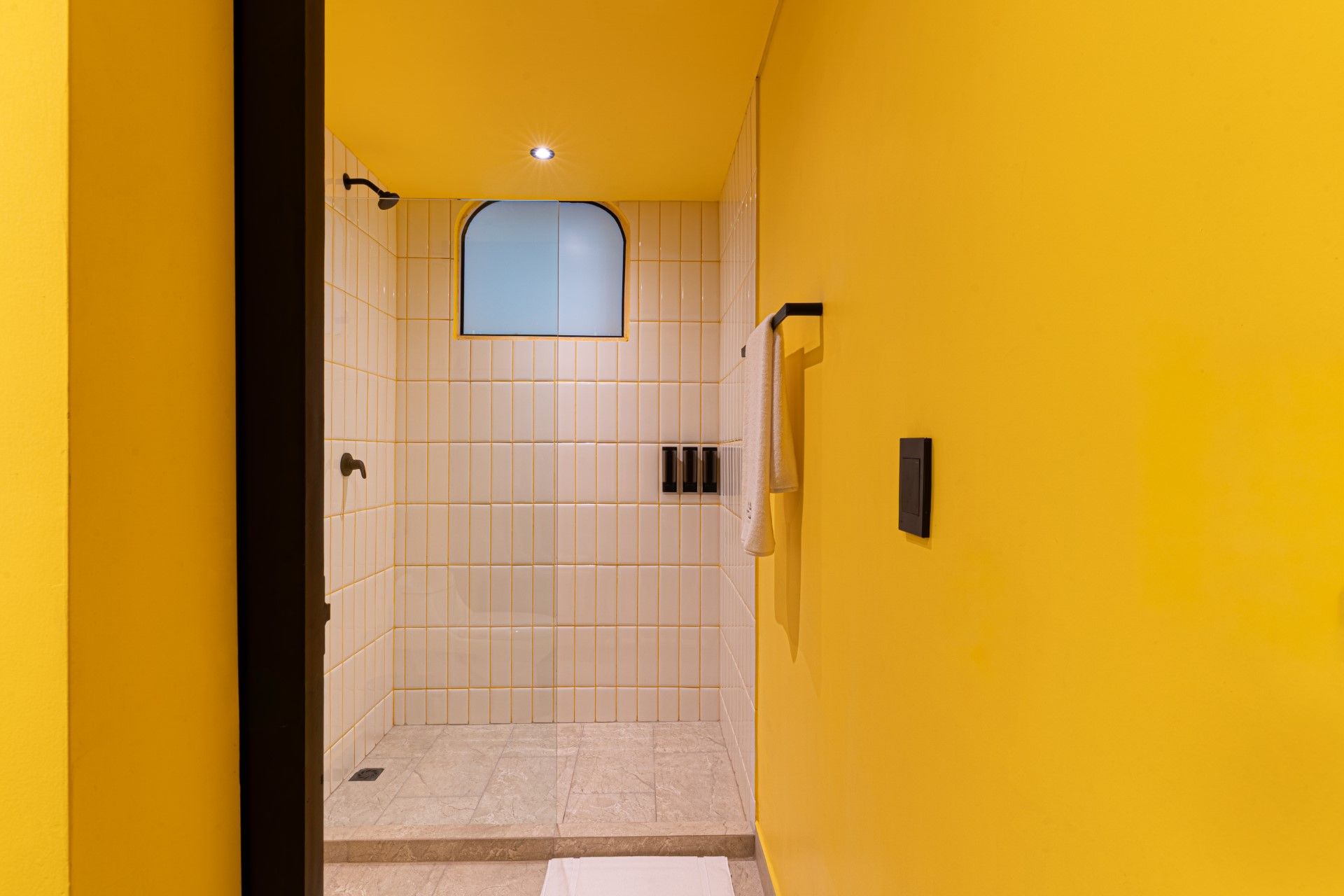 Baño amarillo con ducha. Paredes de azulejos blancos, suelo de terrazo. Ducha negra y toallero.