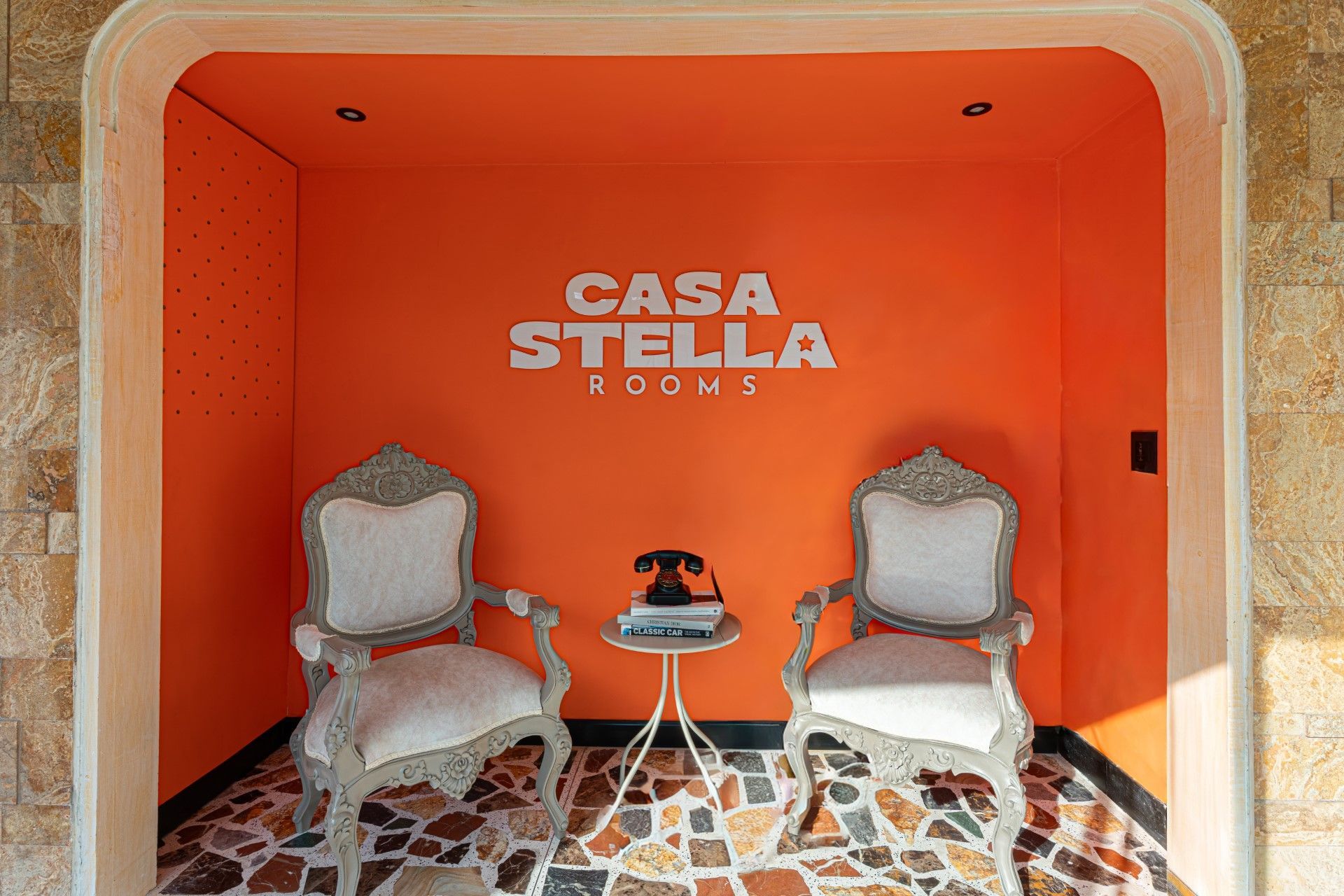 Dos sillas ornamentadas con cojines blancos flanquean una mesita con teléfono, situada en un nicho naranja. 