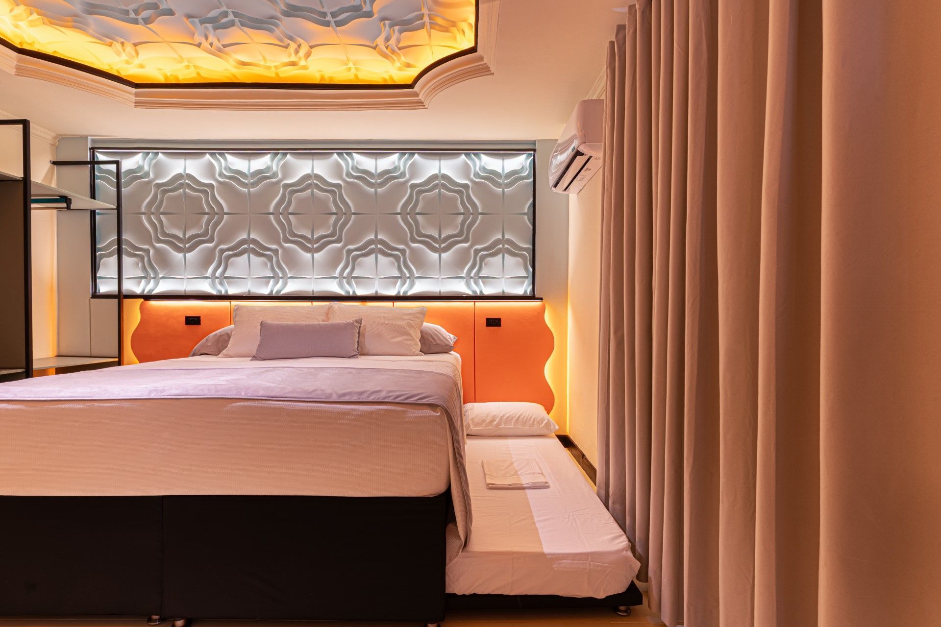 Una habitación de hotel con cabecera naranja, pared estampada, cama, sofá cama y cortinas color crema.