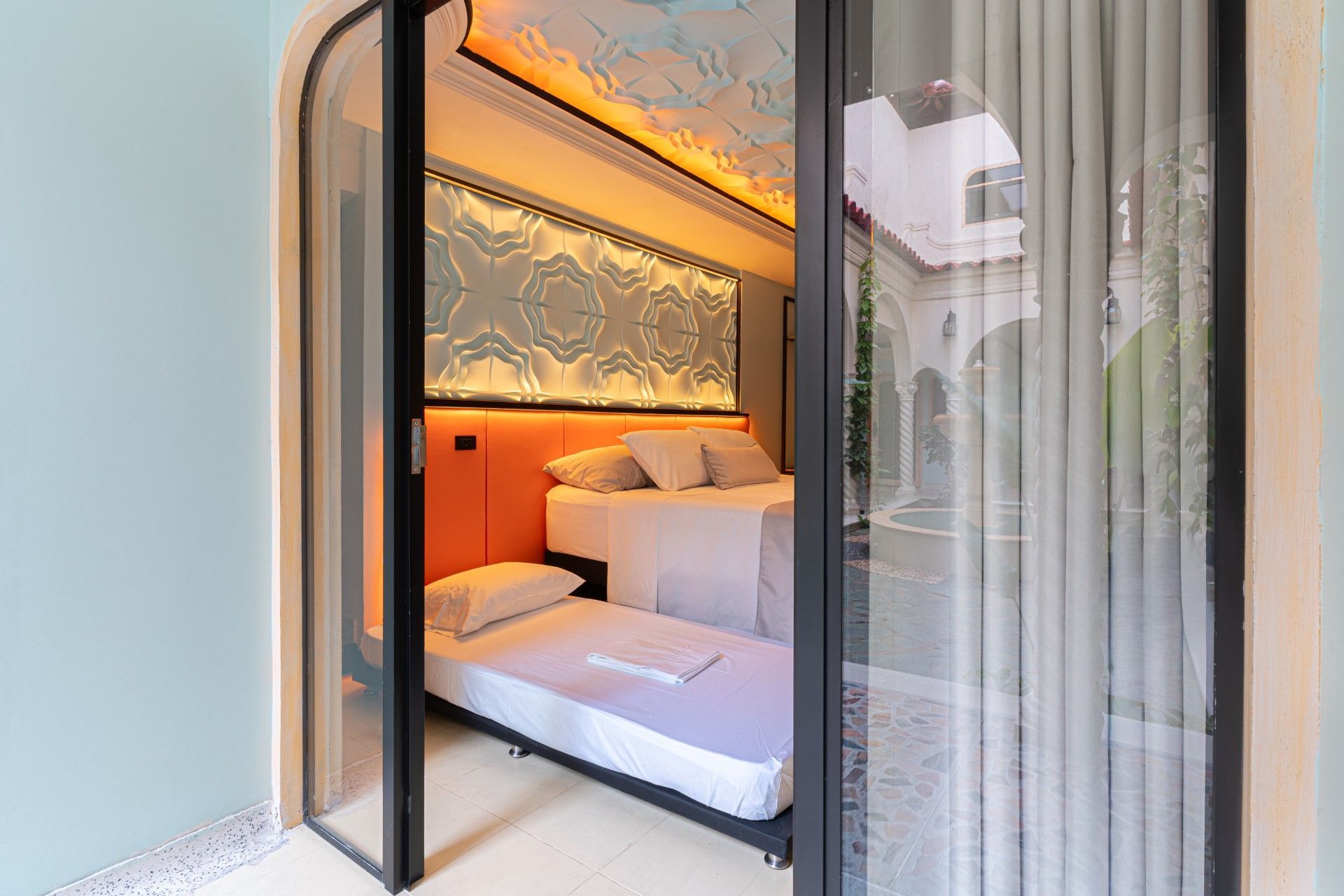 Dormitorio con dos camas, vista a través de puertas de cristal. Cabecero naranja, techo ornamentado y vista al exterior.