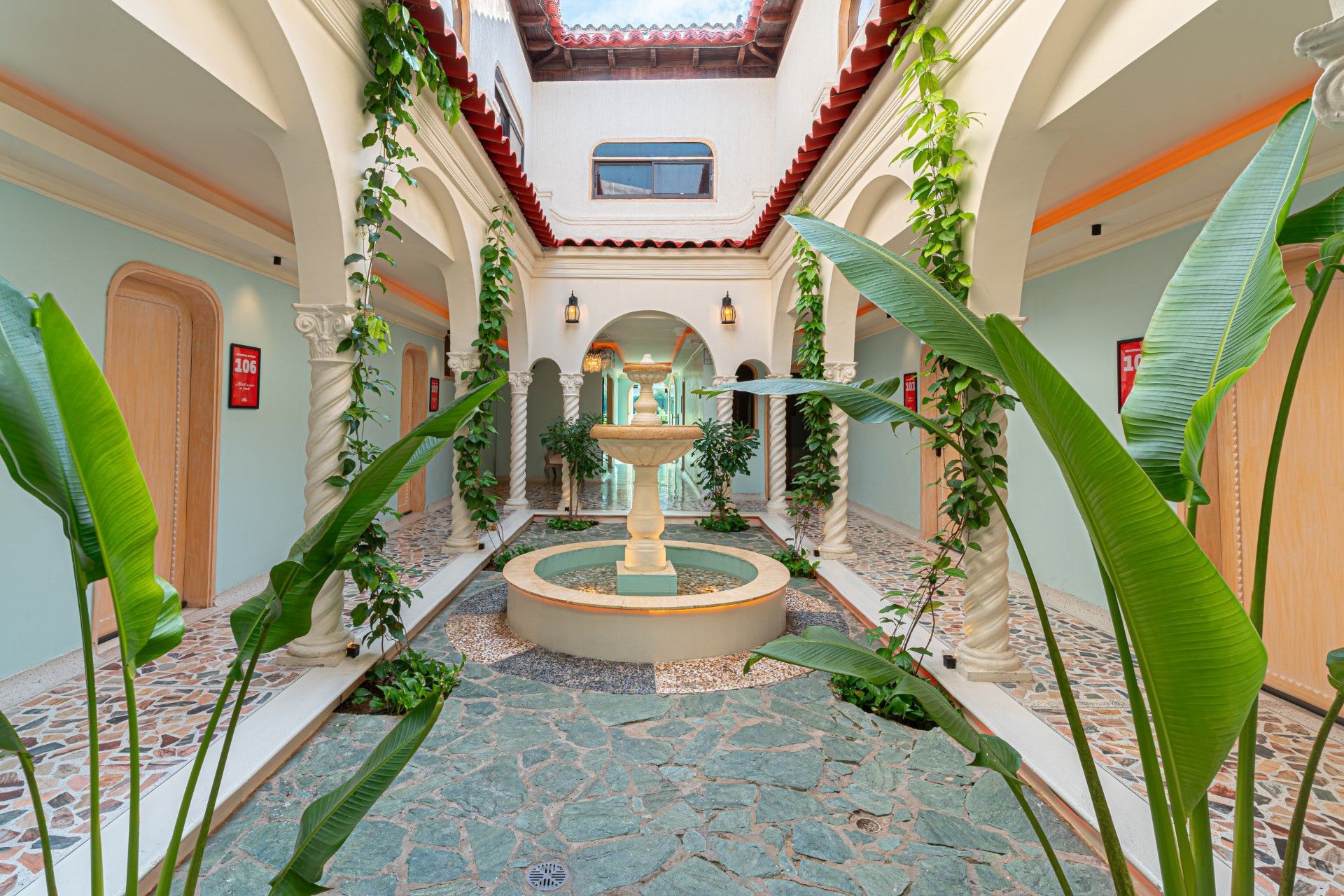 Patio con fuente, arcos y plantas trepadoras. Suelo de baldosas azules, paredes claras y techo abierto.