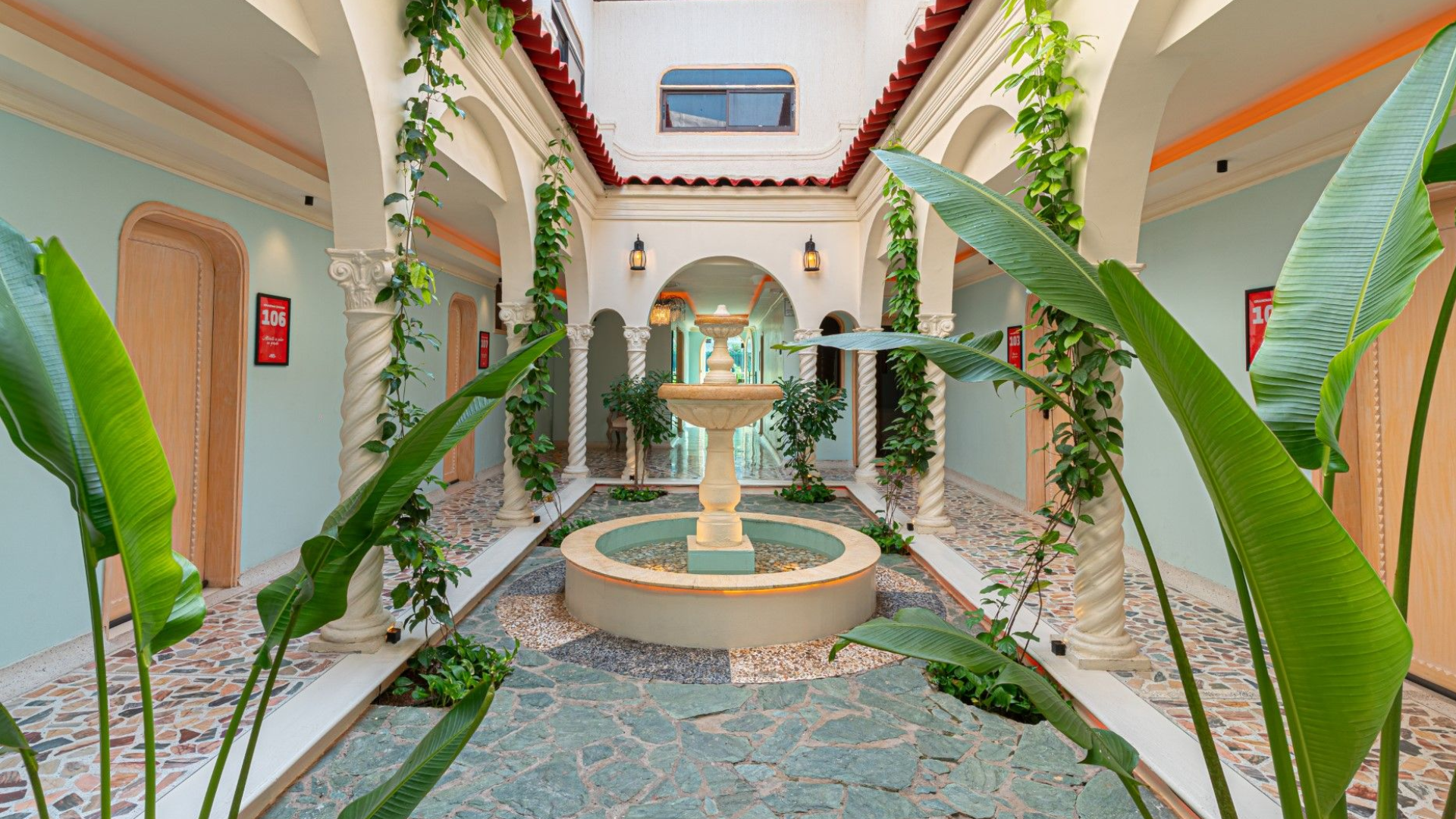 Patio con fuente, arcos y plantas trepadoras. Paredes azul claro, suelo de baldosas y claraboya.