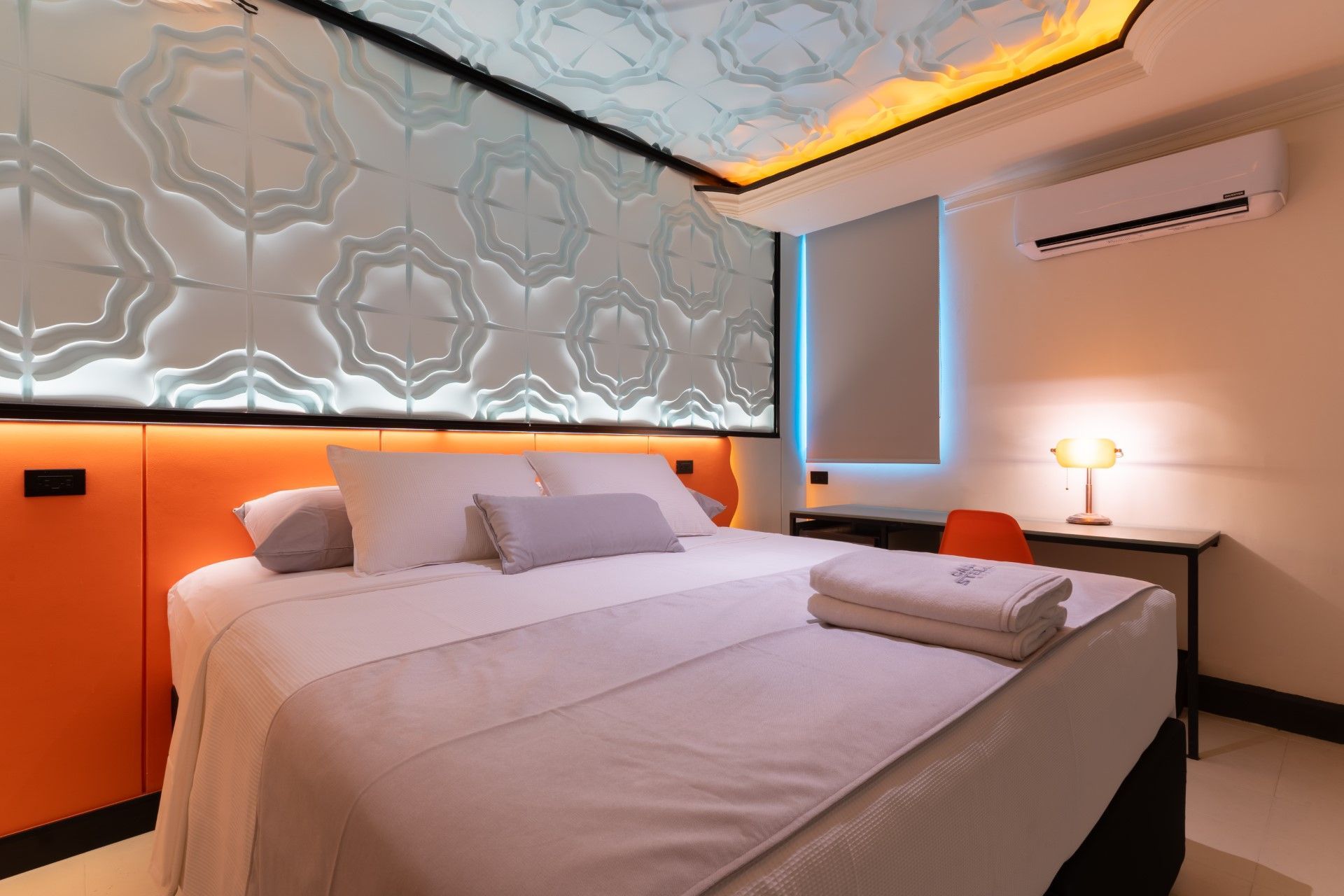 Dormitorio con ropa de cama blanca, detalles en naranja, pared estampada y escritorio.