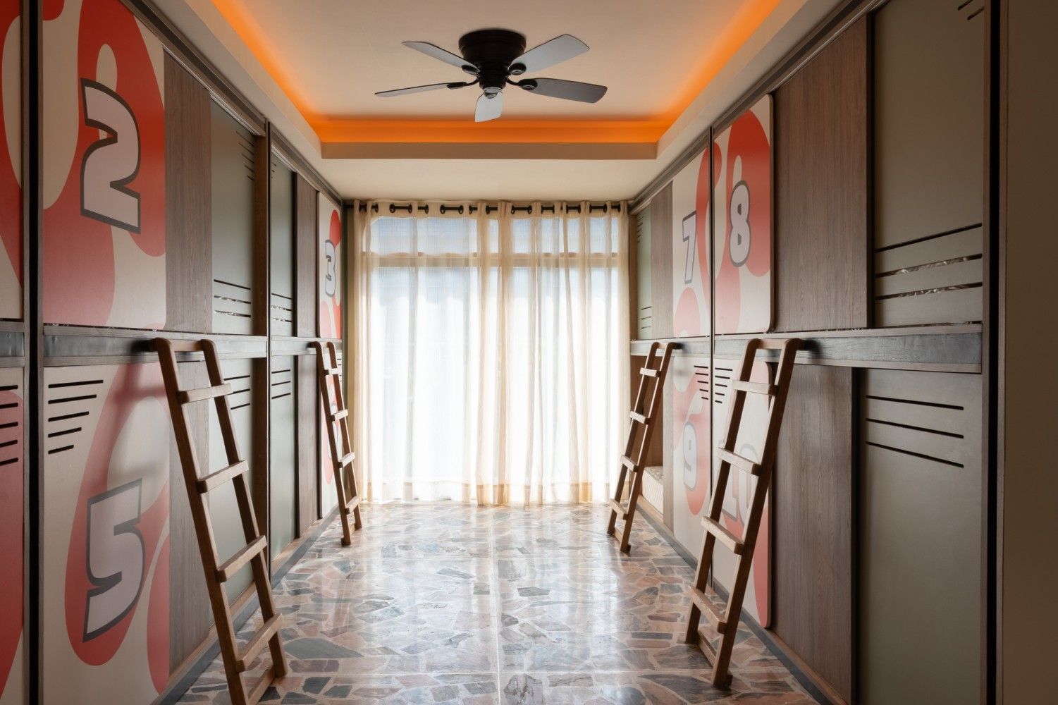 Pasillo estrecho con casilleros numerados, escaleras, ventana con cortinas transparentes y ventilador de techo.