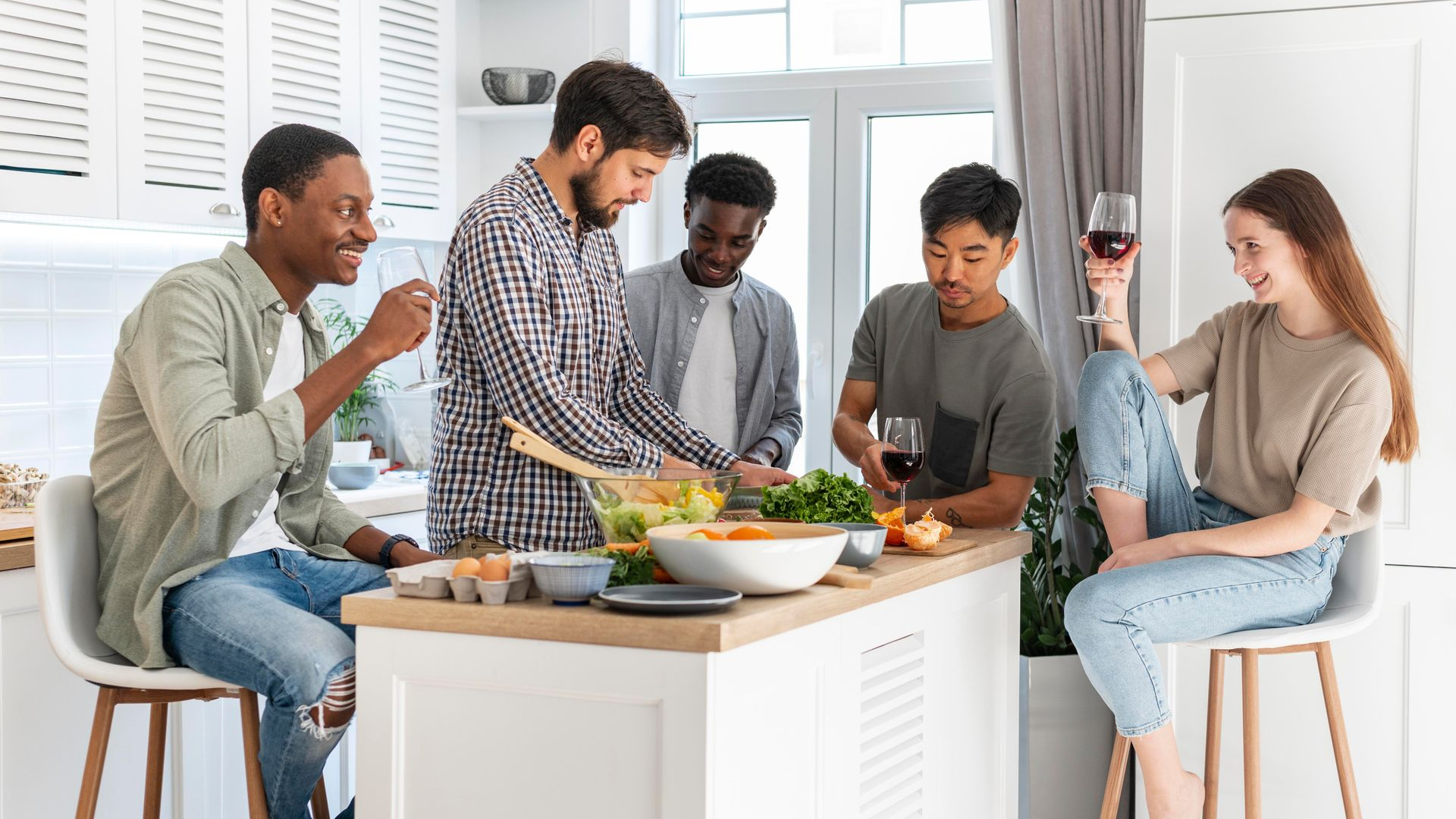Cinco personas en una cocina preparando comida, sonriendo y sosteniendo copas de vino alrededor de una isla de cocina.