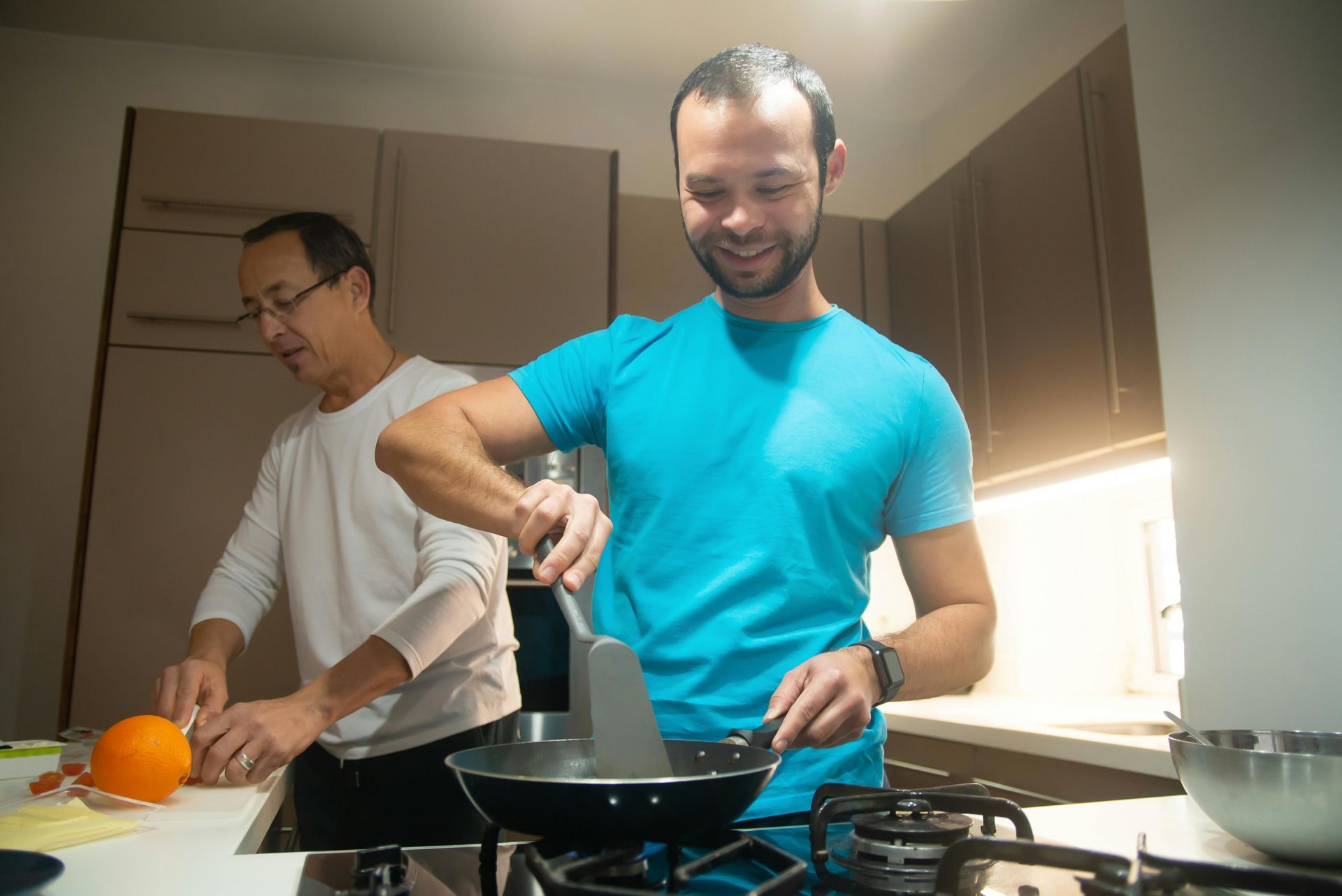 Dos personas cocinando. Una sonríe, usando una espátula en el fuego, mientras la otra pica una naranja.