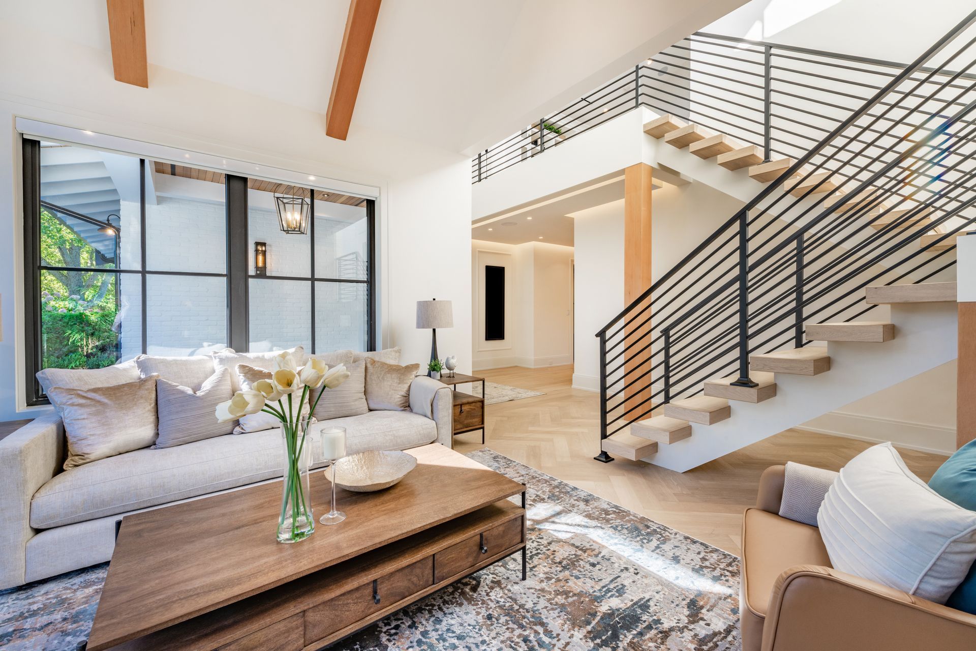 Modern living room with staircase, light wood beams, neutral colors, and large windows.