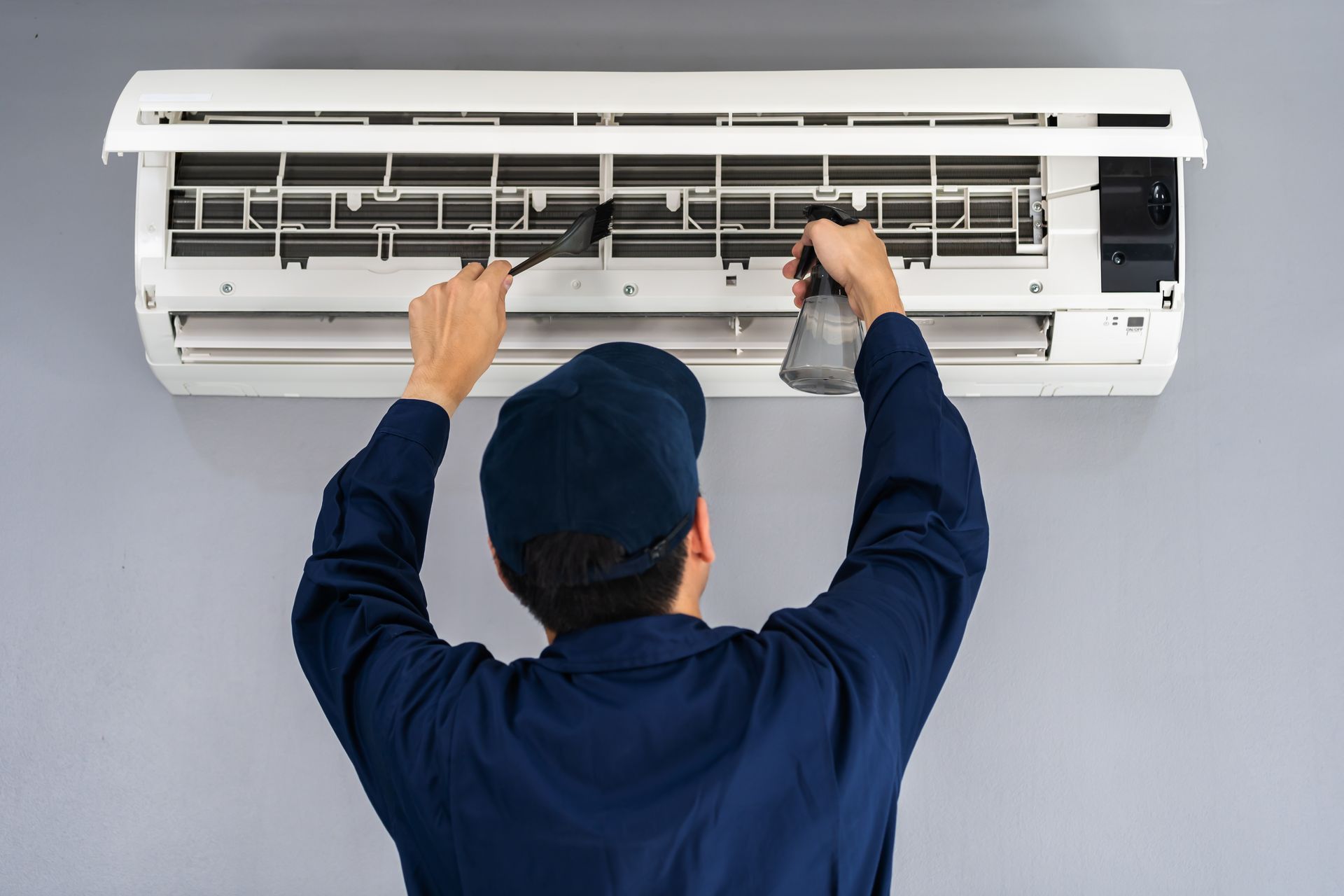 A person in blue uniform cleans a white air conditioner with a brush and spray bottle.
