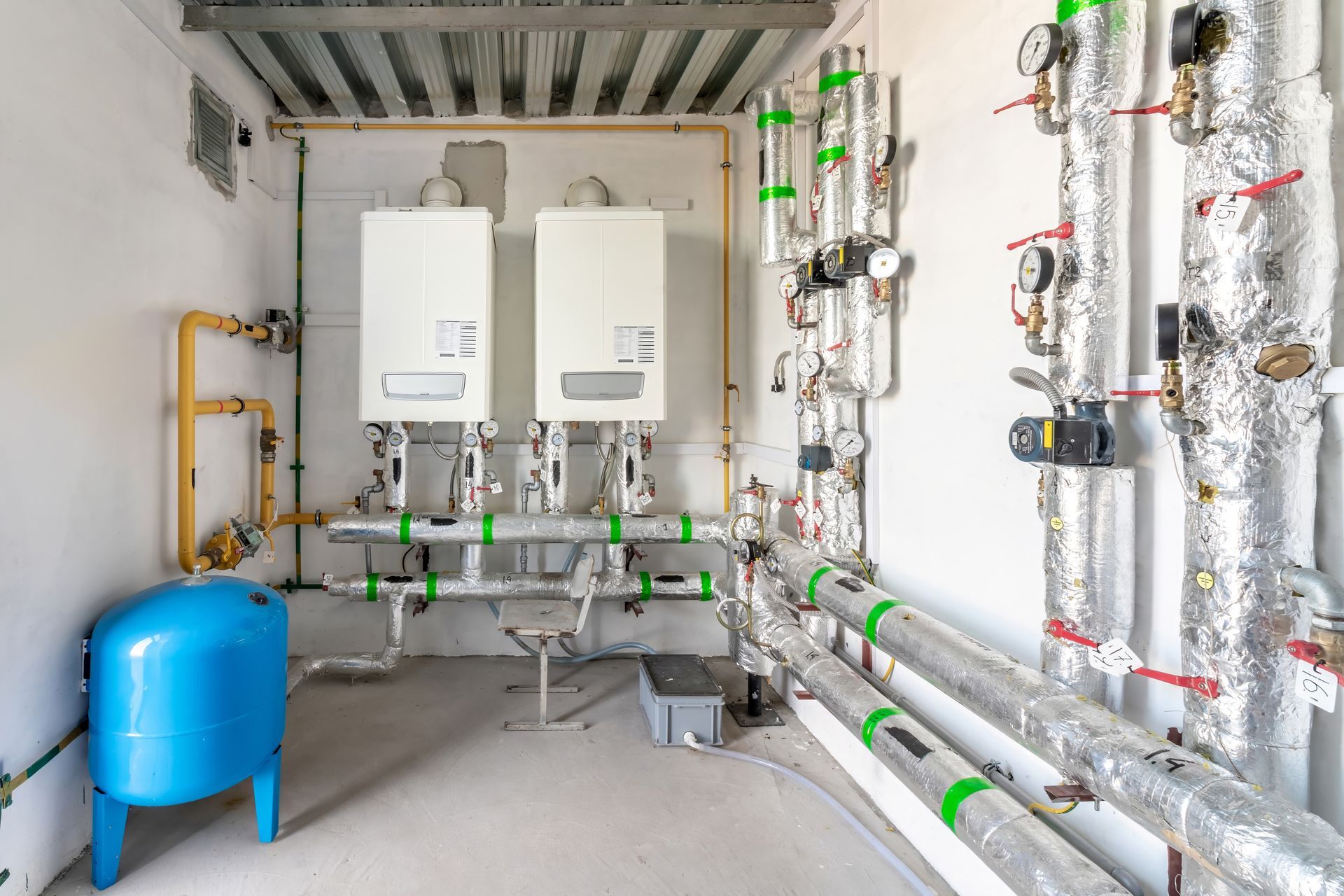 Heating system room with two white boilers, blue tank, and insulated pipes.