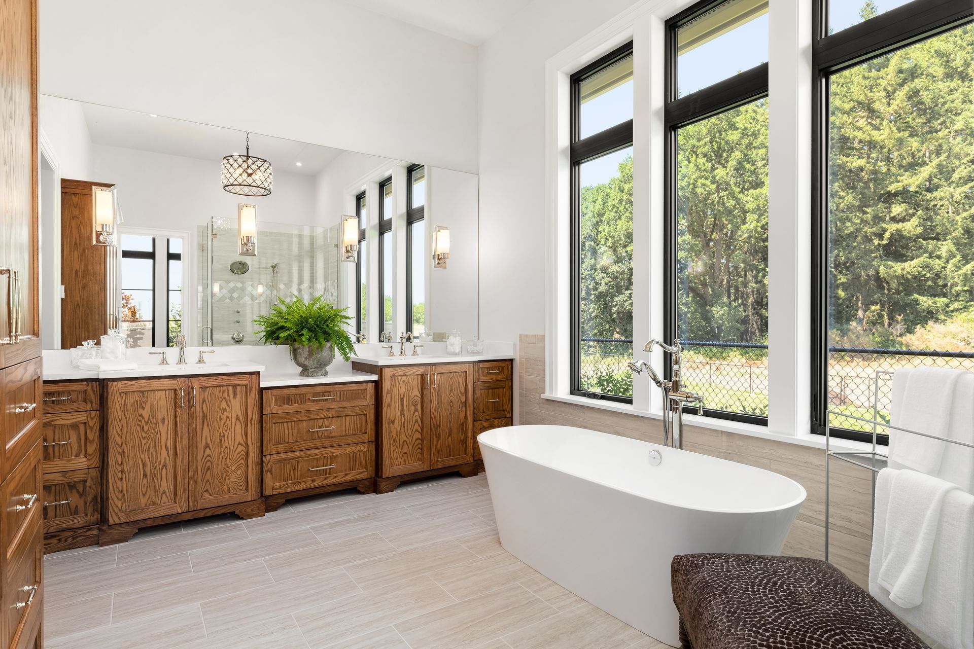 Spacious bathroom with wooden vanities, large mirror, freestanding tub, and tall windows overlooking trees.