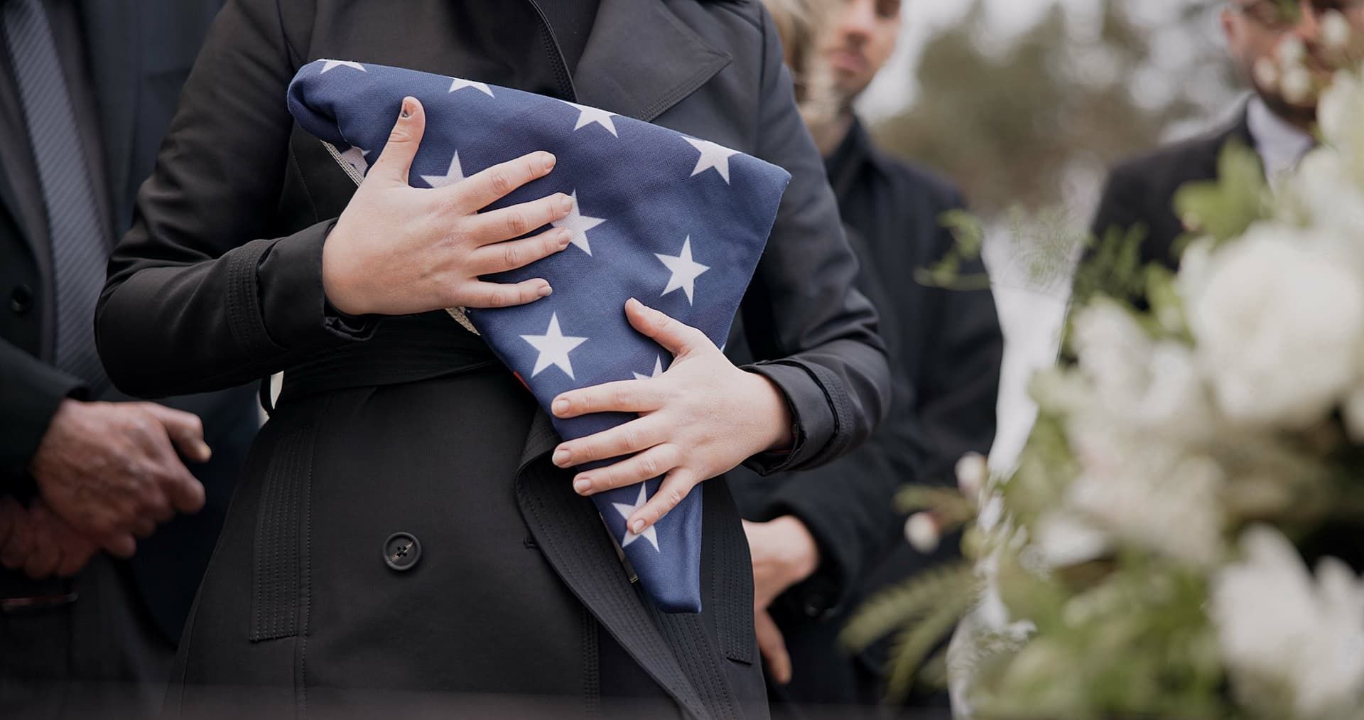 Woman in black coat holds folded American flag at a funeral, surrounded by mourners and white flowers.