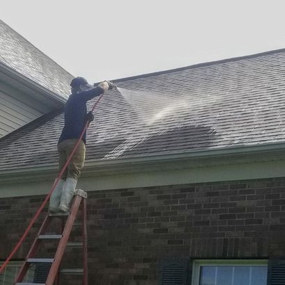 Worker on a ladder pressure-washing a brick house roof, spraying water across the shingles.